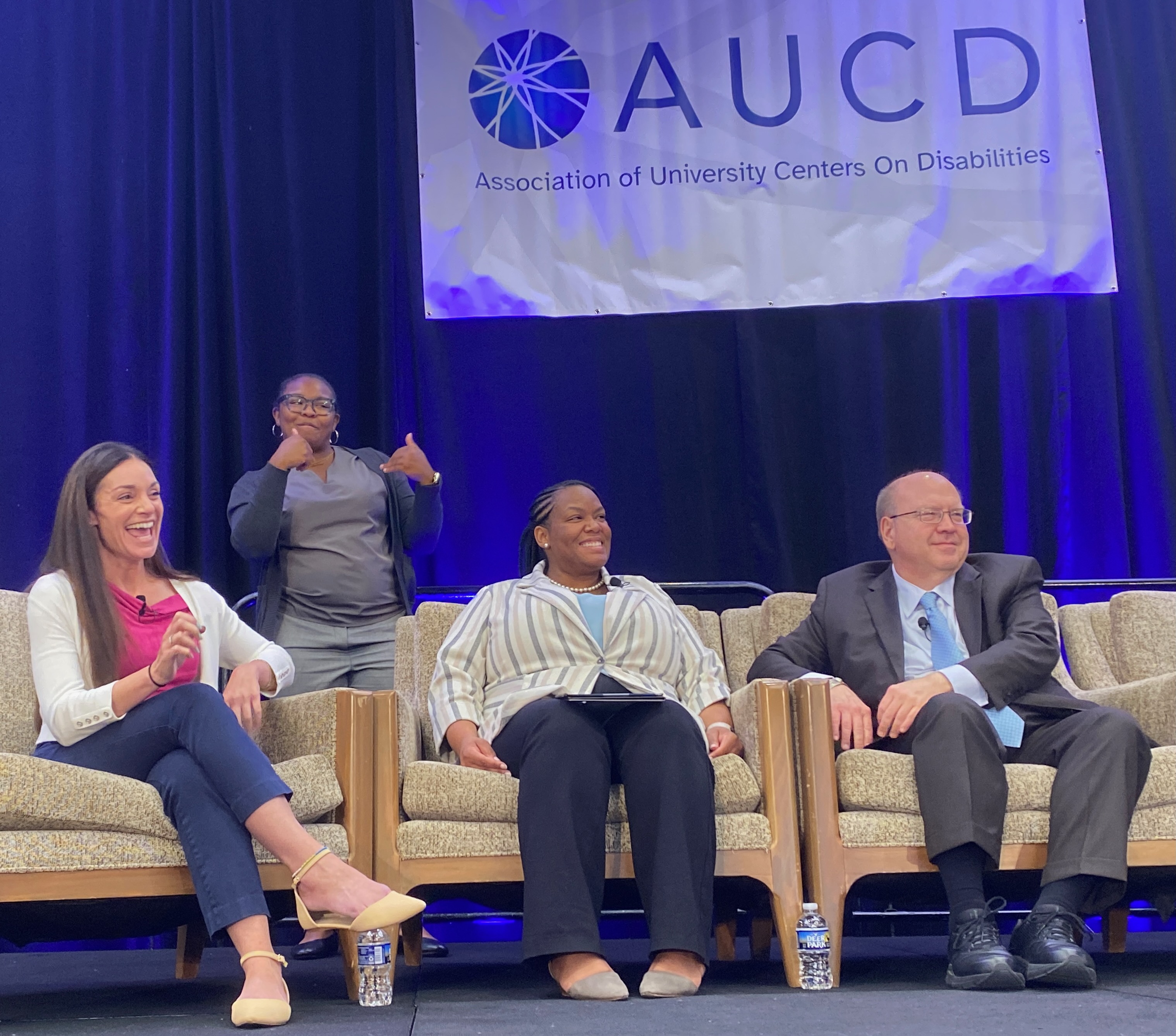 Three panelists from the AUCD 2024 Opening Plenary sit and smile together, an ASL interpreter stands behind them.